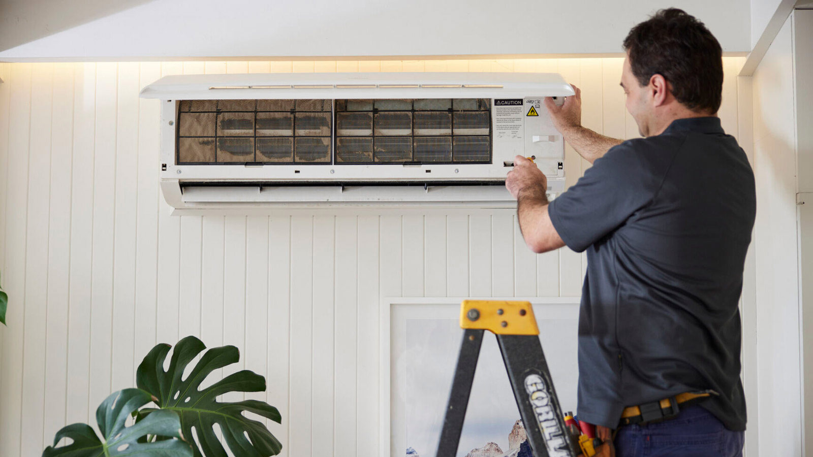 Tradesman standing on a ladder, opening an air conditioning unit and pressing a button.