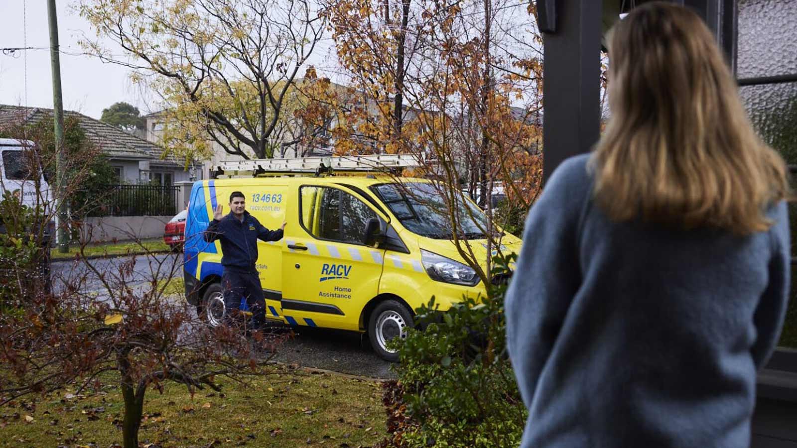 RACV Tradesman in the driveway, holding yellow emergency trades van door open and waving at the woman standing at the front door of a home.