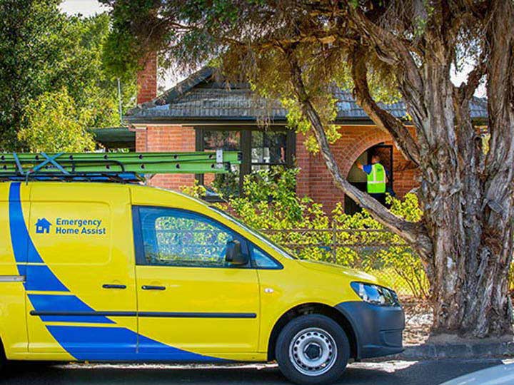 Yellow and blue trades van paked on a street outside of a brick home with tradesman knocking on the front door.