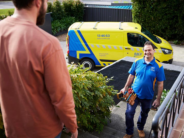 RACV Tradesman smiling and greeting homeowner on front porch with yellow and blue tradie van in background.