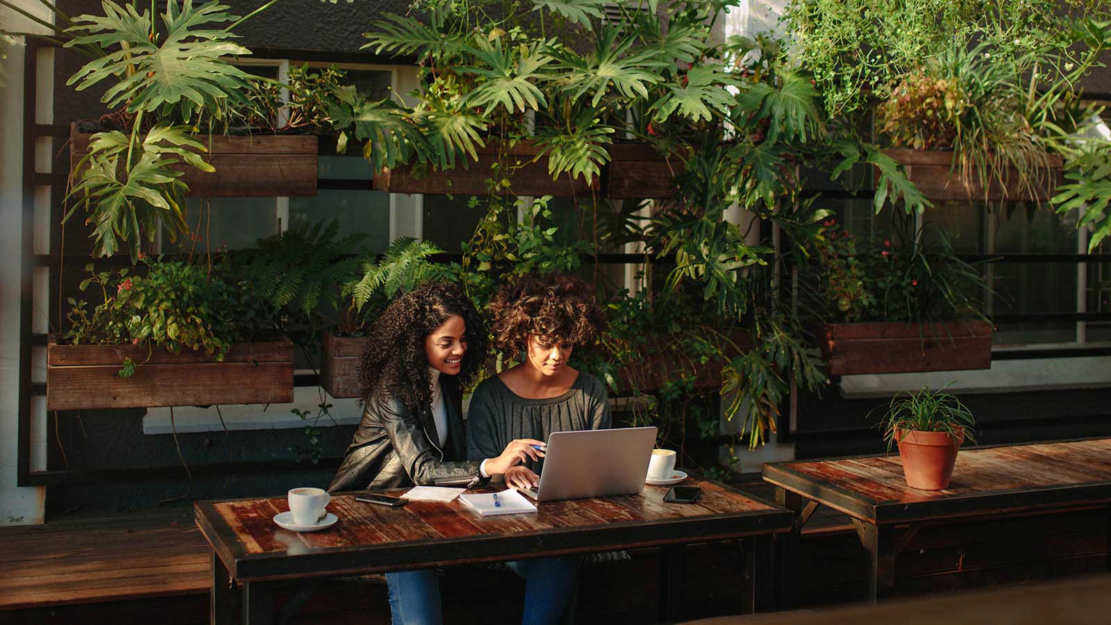 Two woman sitting in a stylish outdoor courtyard drinking coffee and looking at a laptop.