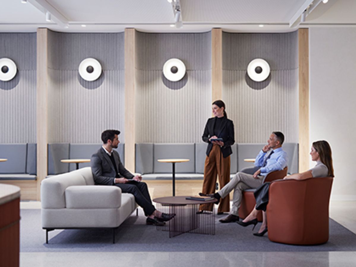 Four people in busines clothes having a meeting in an open cowork space. One person is standing and holding a tablet, the other three people are sitting on couches around a coffee table.
