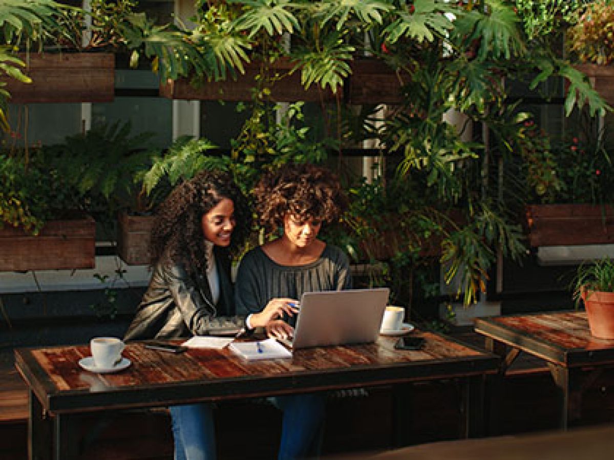 Two woman sitting in a stylish outdoor courtyard drinking coffee and looking at a laptop.