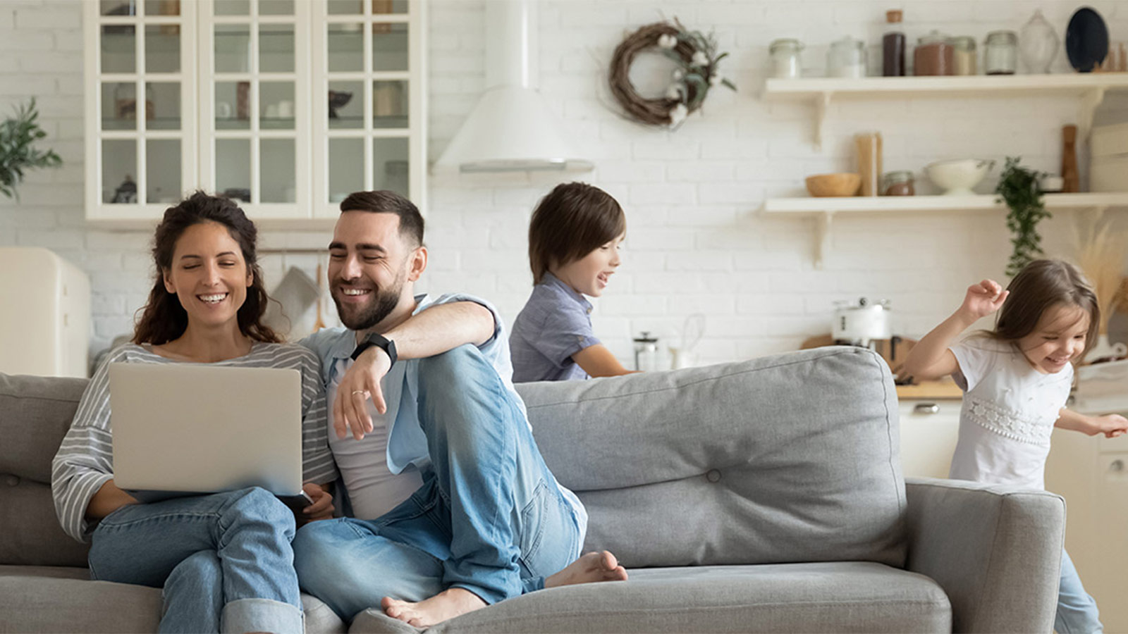 Parents sitting on the couch smiling at their laptop while their kids are playing together.