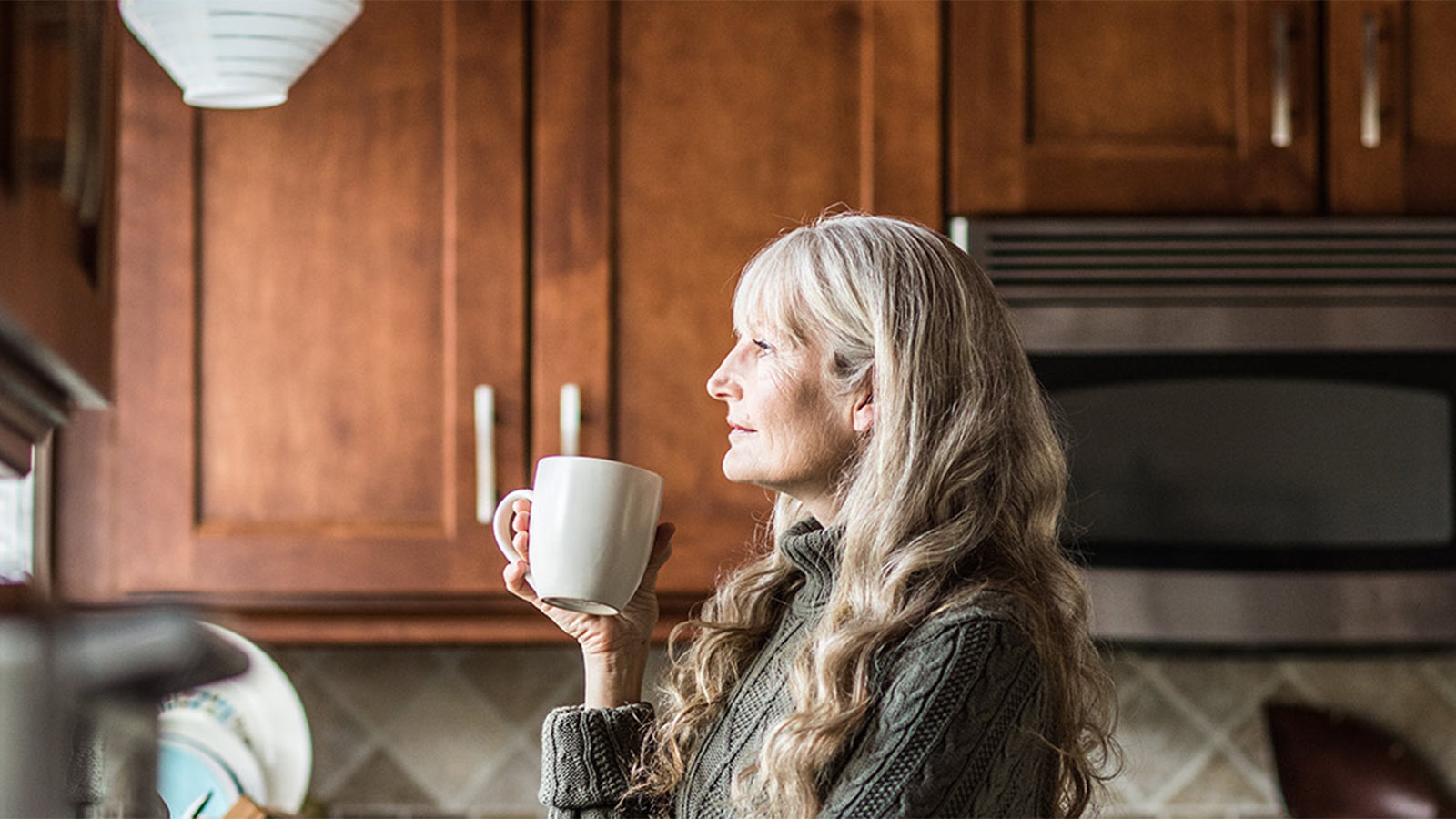 Woman standing in her kitchen holding a white mug and staring out the window.