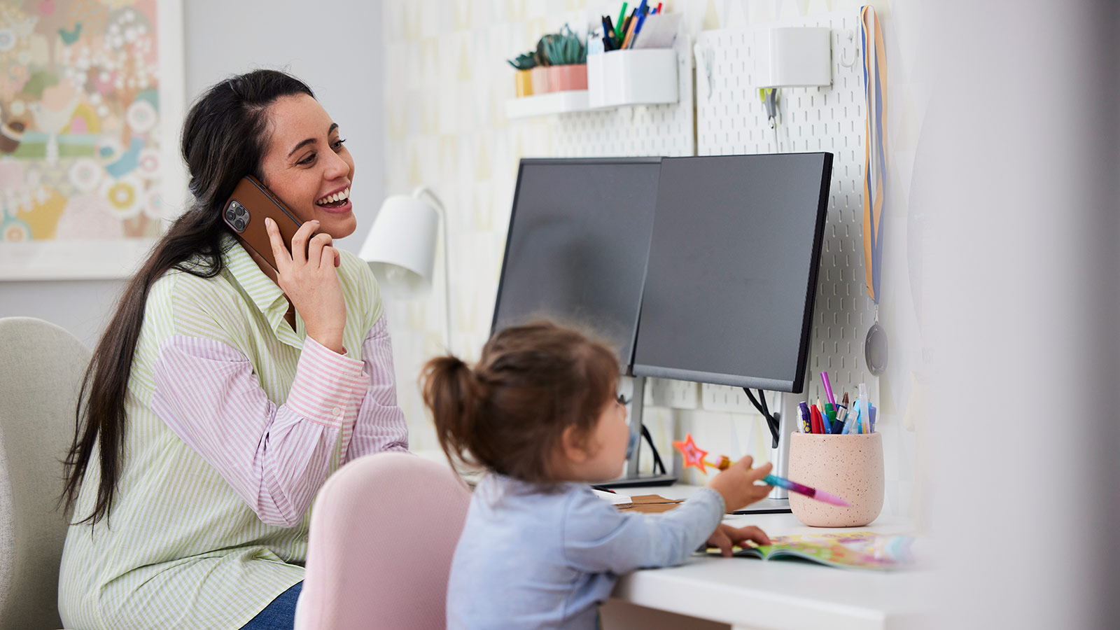 Parent laughing on the phone and young child drawing while sitting at a desk together.