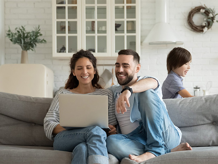 Parents sitting on the couch smiling at their laptop while their kids are playing together.