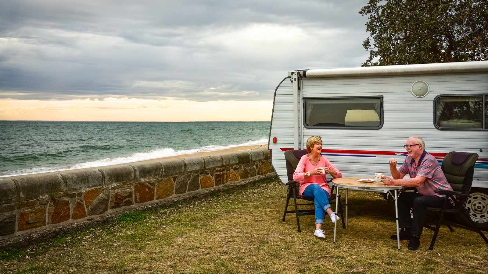 Elderly couple drinking tea and laughing in front of their caravan, parked next to the beach.