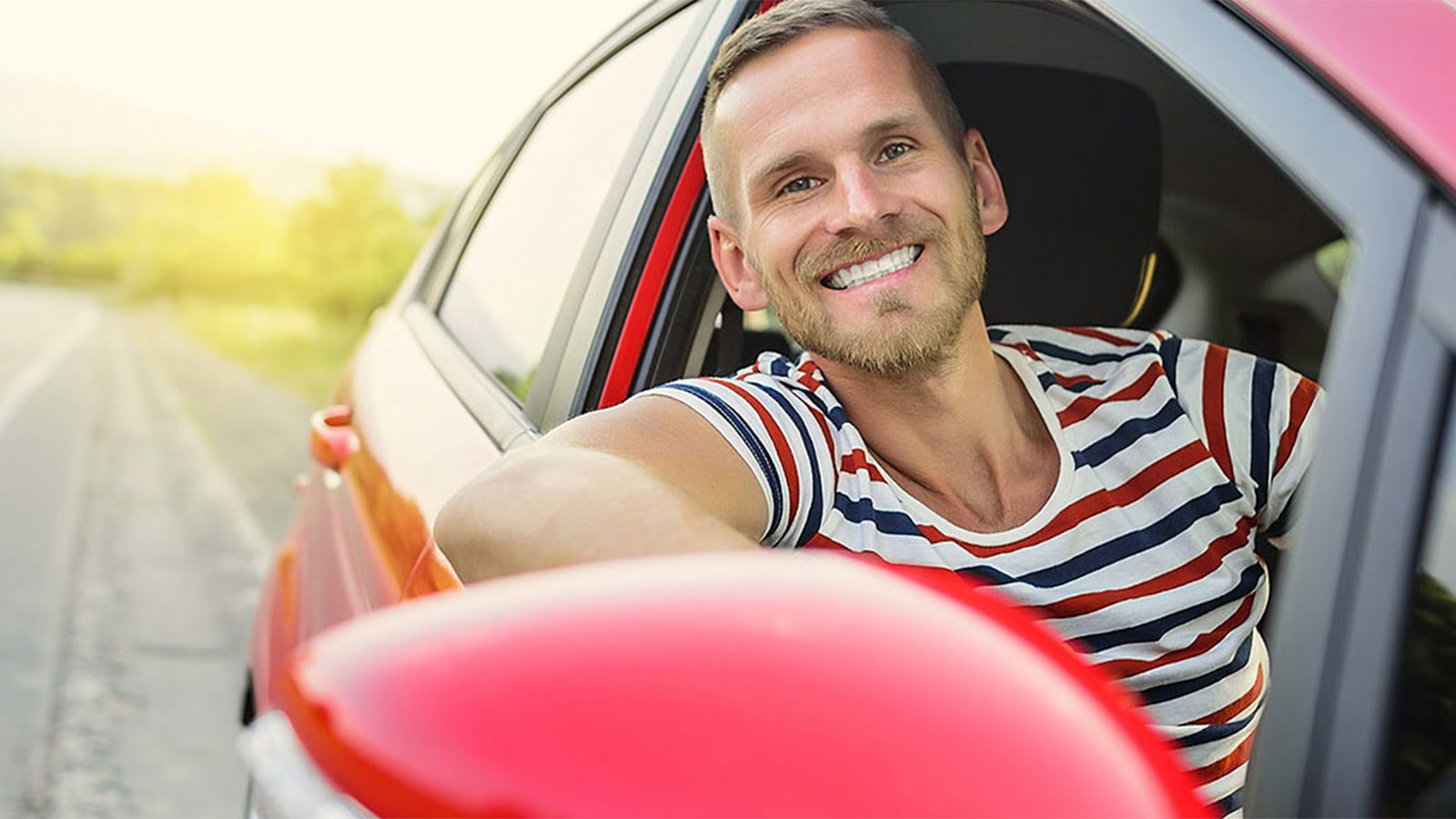 Man sitting in the drivers seat of a red car and smiling out the window.