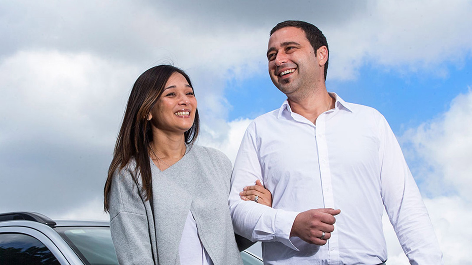 Young couple linking arms and smiling outside, in front of their silver car.