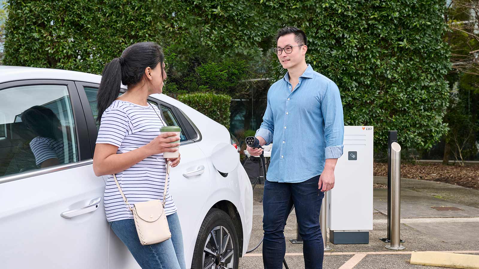 Two people standing near a white electrical vehicle at a charging station. One person is holding a charger, and the other is holding a takeaway coffee mug.