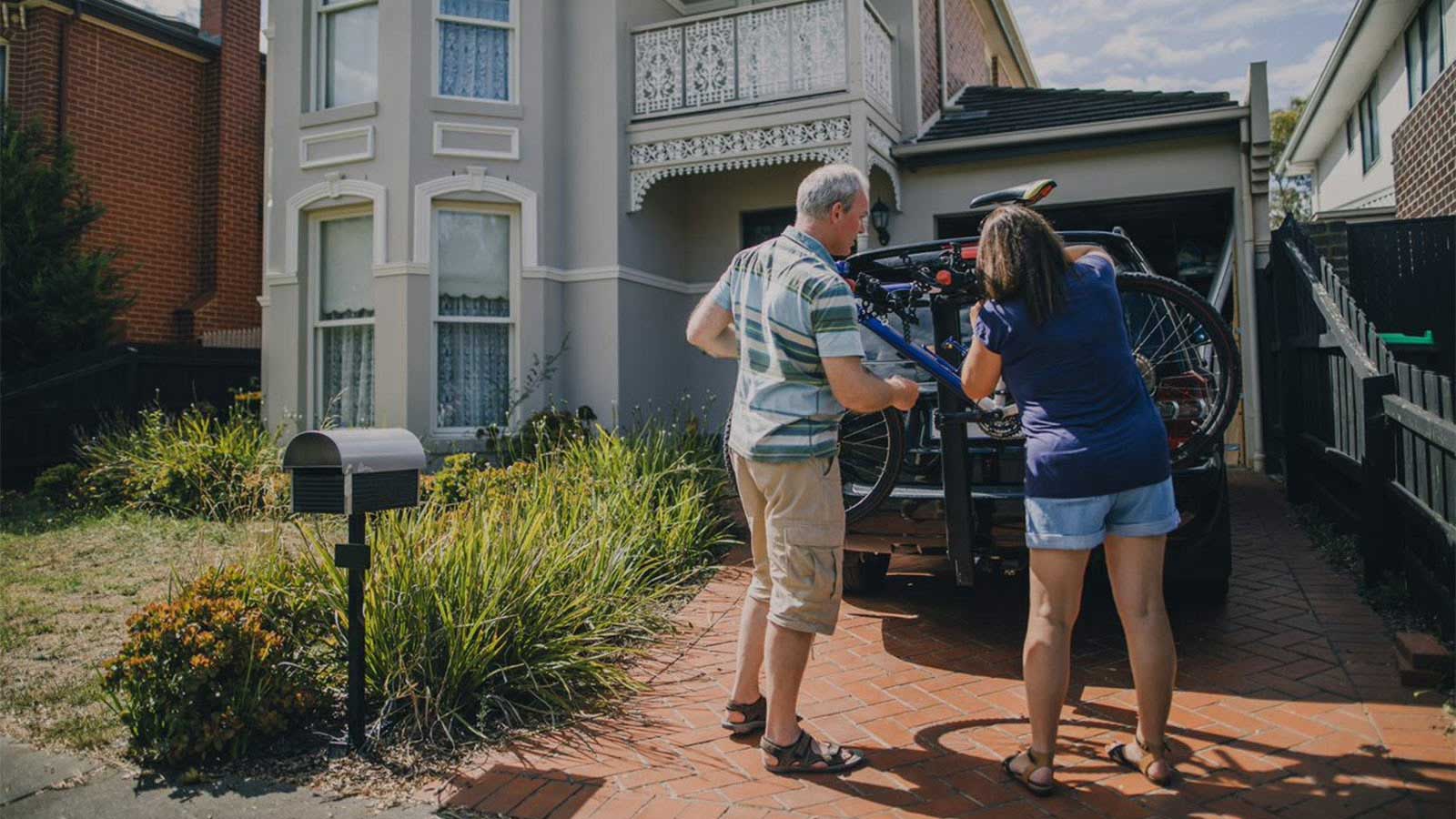 Young woman helping elderly man affix a bicycle to a car parked on a brick driveway.