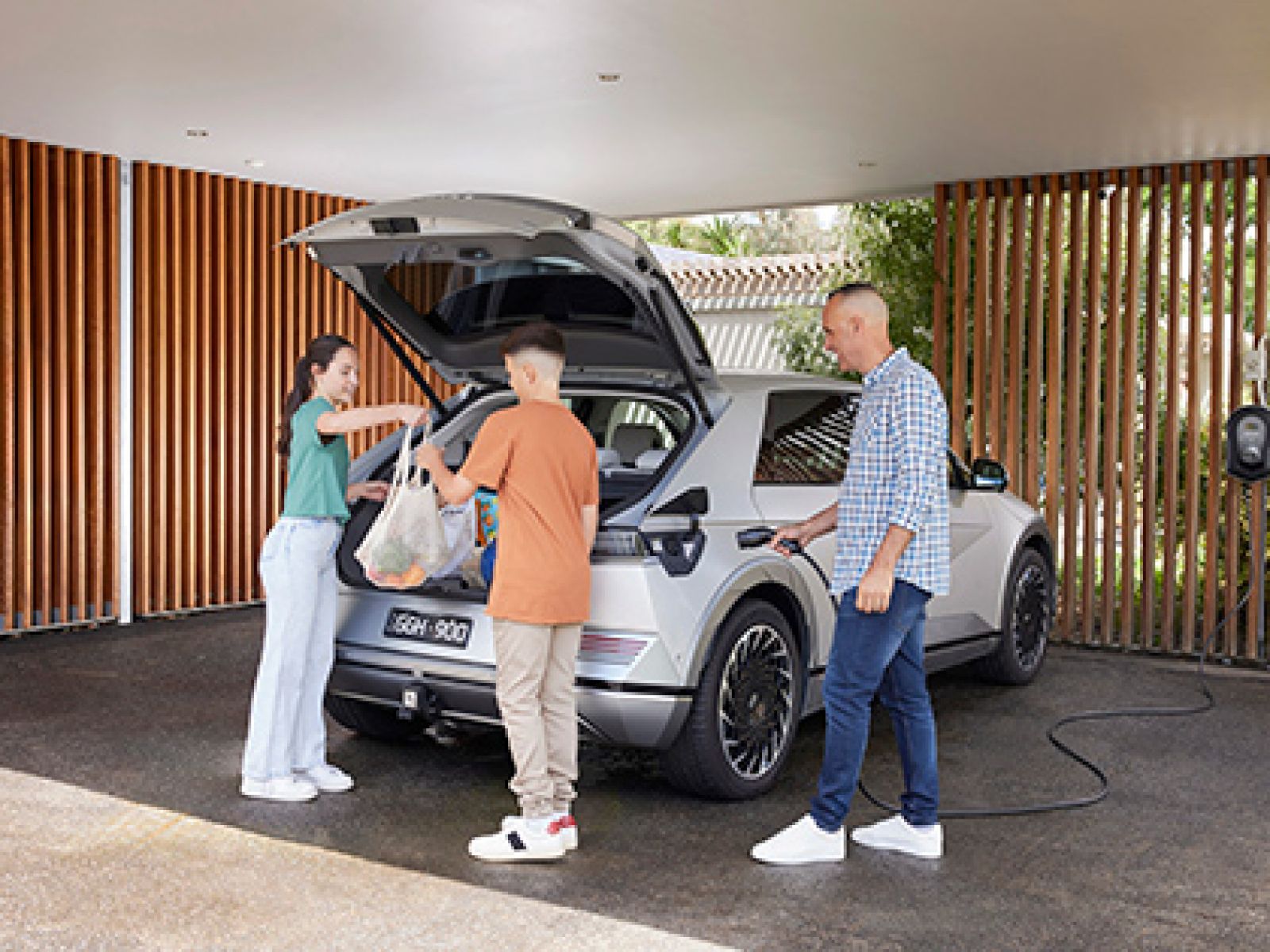 Parent charging a white electric SUV in a carport, while two kids unload groceries from the boot.