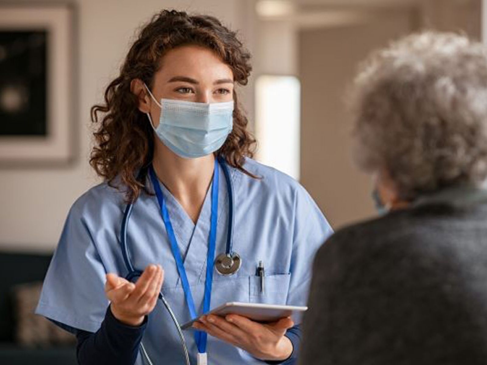 Nurse wearing a mask and scrubs talking to an elderly woman.