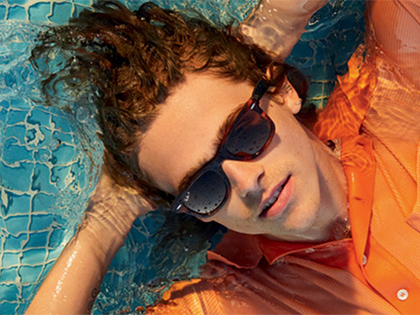 Man in an orange polo lying in a swimming pool, wearing black sunglasses.