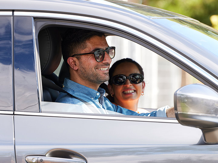 Two people wearing sunglasses, sitting in the front seats of a car and smiling together.