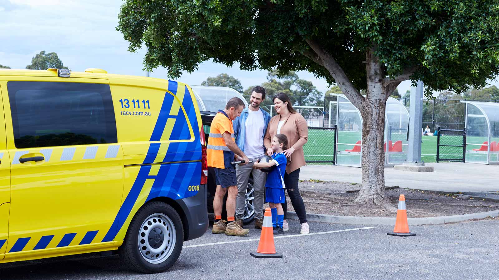 Smiling young couple and child speaking with an RACV roadside assistance mechanic near a soccer field.
