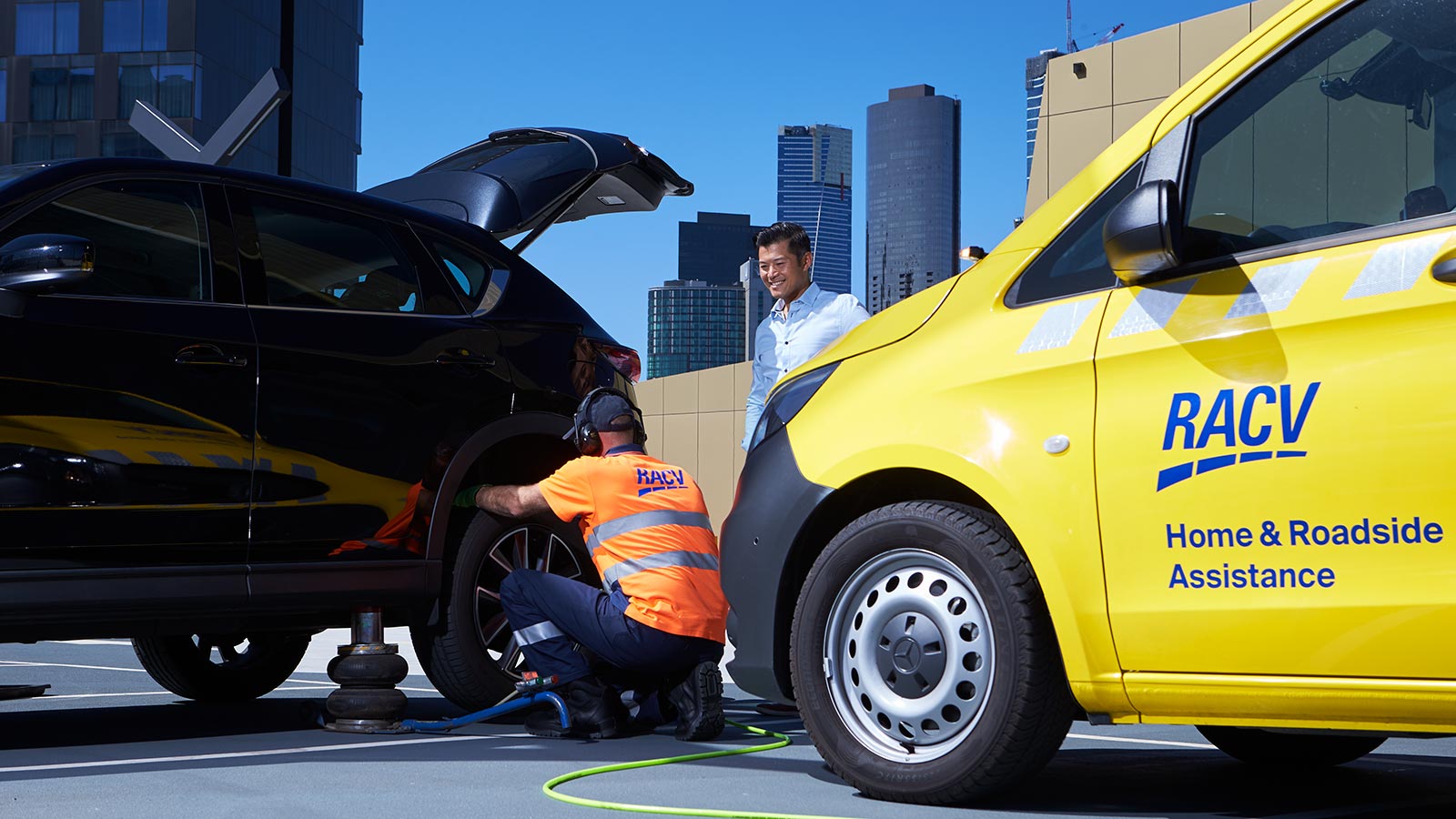 An RACV Emergency Roadside Assistance expert in high-vis kneeling down and changing the back left wheel of a black SUV. A person in business clothes stands next to them, and a yellow RACV Home and Roadside Assistance van is parked nearby.