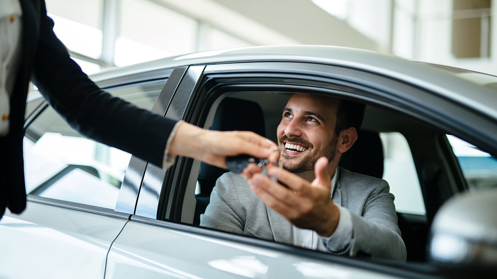 Person in business clothes sitting in the driver's seat of a silver sedan at a car dealership, smiling as someone passes them keys through the window.