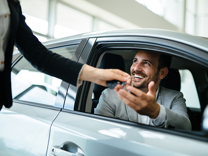 Person in business clothes sitting in the driver's seat of a silver sedan at a car dealership, smiling as someone passes them keys through the window.