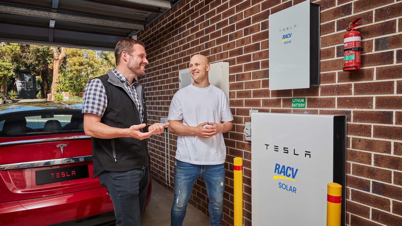 Two smiling men talking in front of a red Tesla and Tesla powerwall battery.