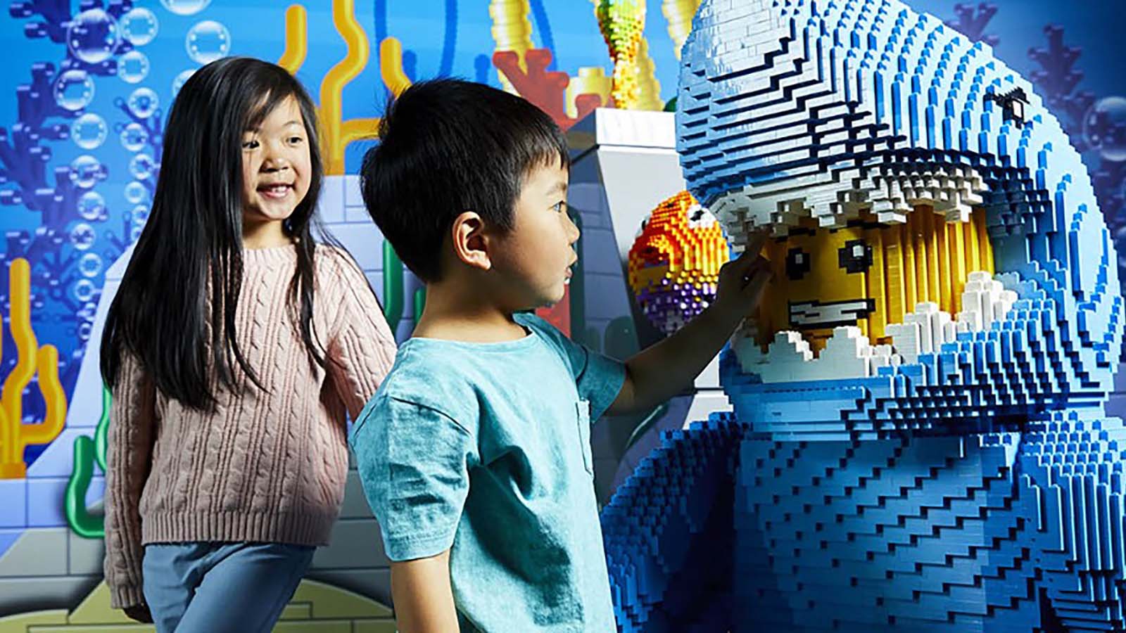 Two children looking at statue of a man in a whale suit made out of lego.