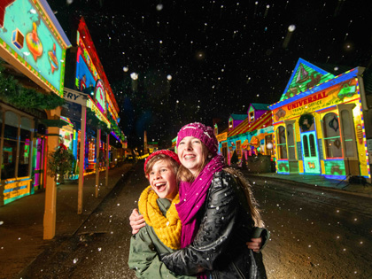 Two children gugging with huge smiles standing in the snow surrounded by colourful lit up buildings at Sovereign Hill.