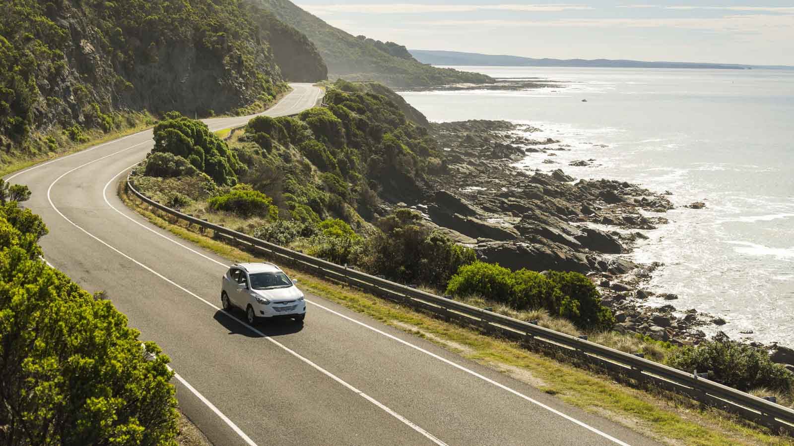 A white SUV driving along a costal road on a sunny day. 