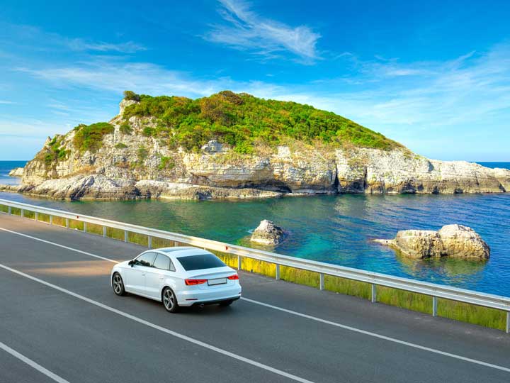 White car driving along a scenic costal road with a large island rock in the water nearby.