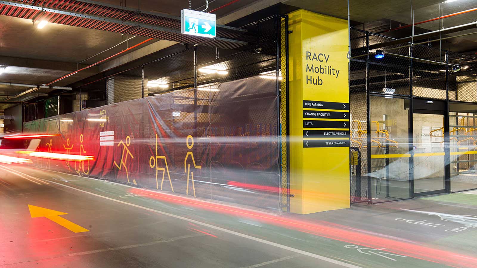 Bright lights created from moving vehicles in the underground Mobility Hub with a large yellow signs showing directions for bike parking, change facilities, lifts and electric vehicle charging.