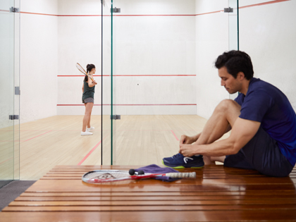 Man tying his shoe whilst seated in front of a woman playing sqaush behind galss doors.