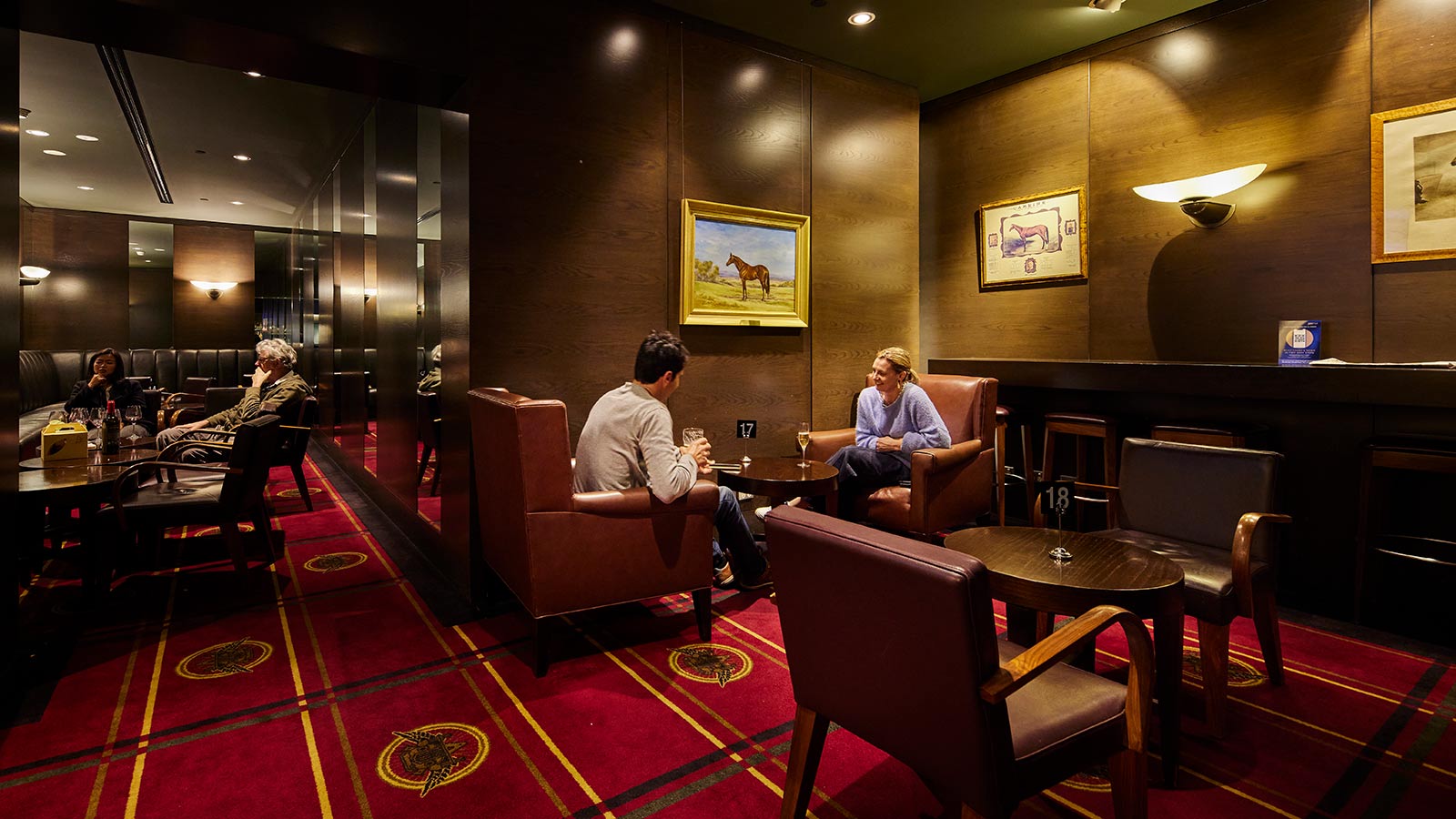 People sitting together with drinks at the Carbine Bar at RACV City Club, featuring leather chairs around wooden side tables, low lighting and red carpet.