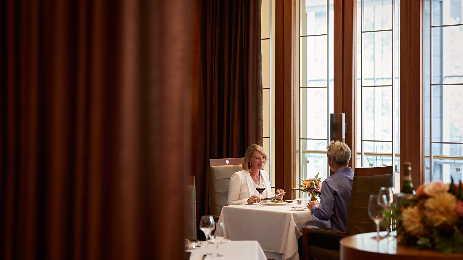 A couple dining at a table covered with a white tablecloth, next to a window at RACV City Club's Members Lounge.