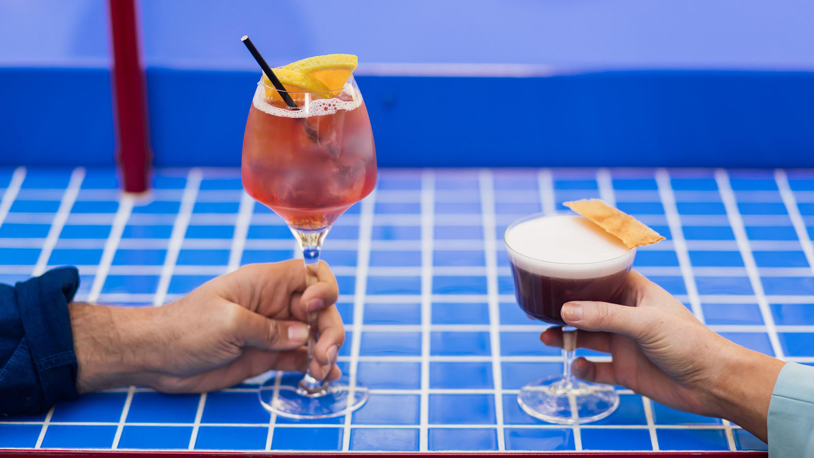Two hands holding cocktails over the blue-tiled bar at Sojourn.