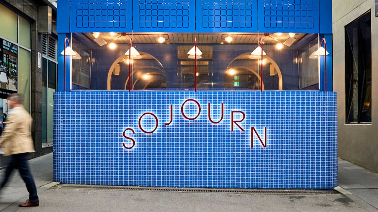 An image of a man strolling past Sojourn, a structure covered in blue tiles with the name in red on the side.