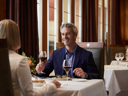A man wearing a blue suit with a glass of red wine smiling at a woman seated at a table. 