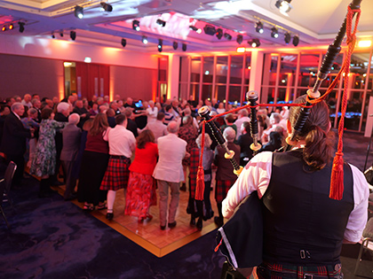Large group of people folk dancing on the dancefloor of a function room, while a person plays the bagpipes nearby.