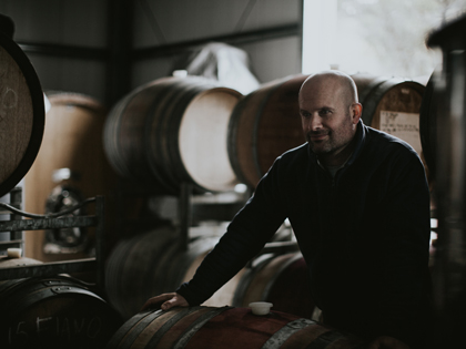 A man in a wine cellar leaning on a wine barrell.