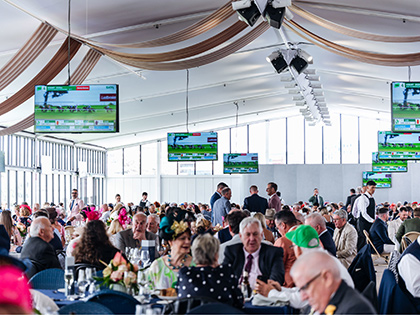 Crowd of people sitting at tables in a marquee with televisions showing a horse race.