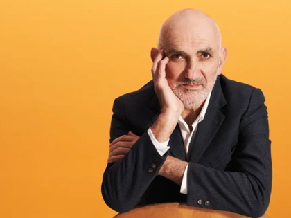 Paul Kelly posing with his hand on his face looking at the camera infront of an orange background.