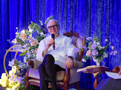 A man with a microphone in a seat surrounded with florals. 