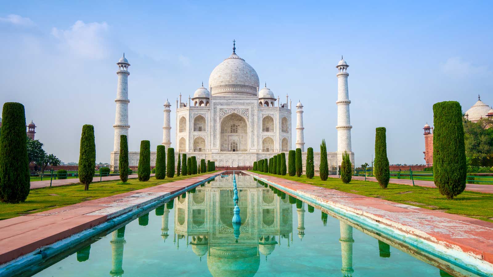 A beautiful white building in India with its reflection shinign in the water lined with trees. 