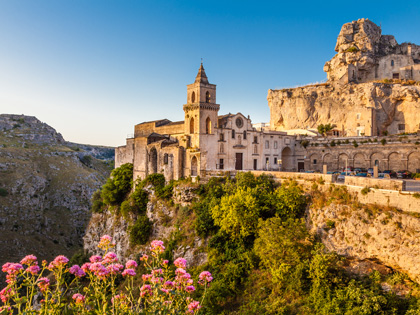 A castle on cliffs glowing in the sunlight surrounded by grenery and pink flowers. 