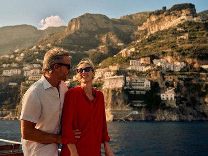 A couple smiling at each other whilst on a boat with a beautiful European town behind them