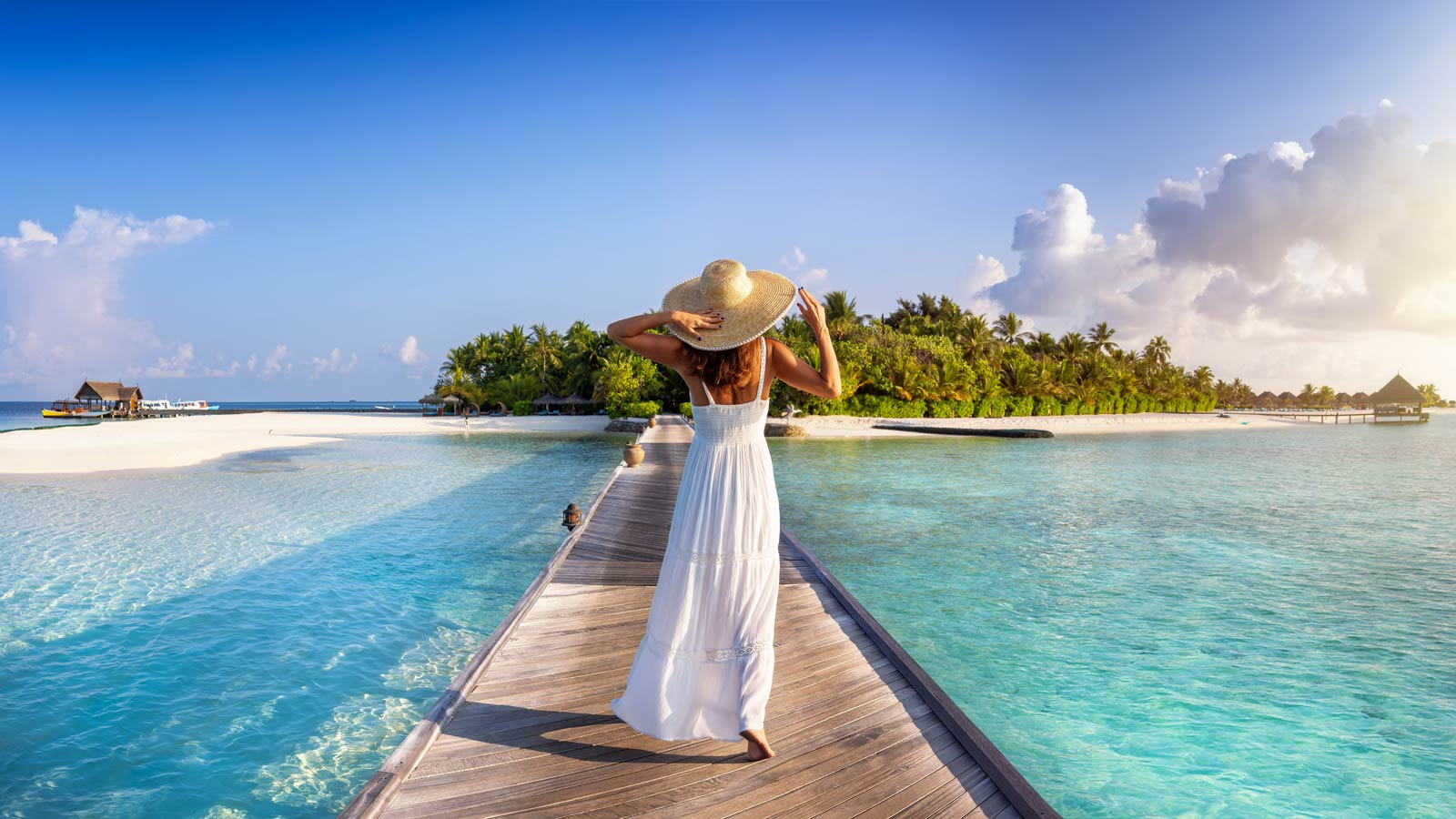 Woman in a white sun dress and large sun hat standing at the eand of a pier surrounded by crystal clear water looking back at a tropical island.