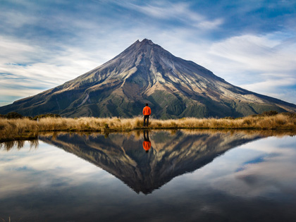 Man standing infront of Mount Taranaki in New Zealand with the reflection showing in the lake.