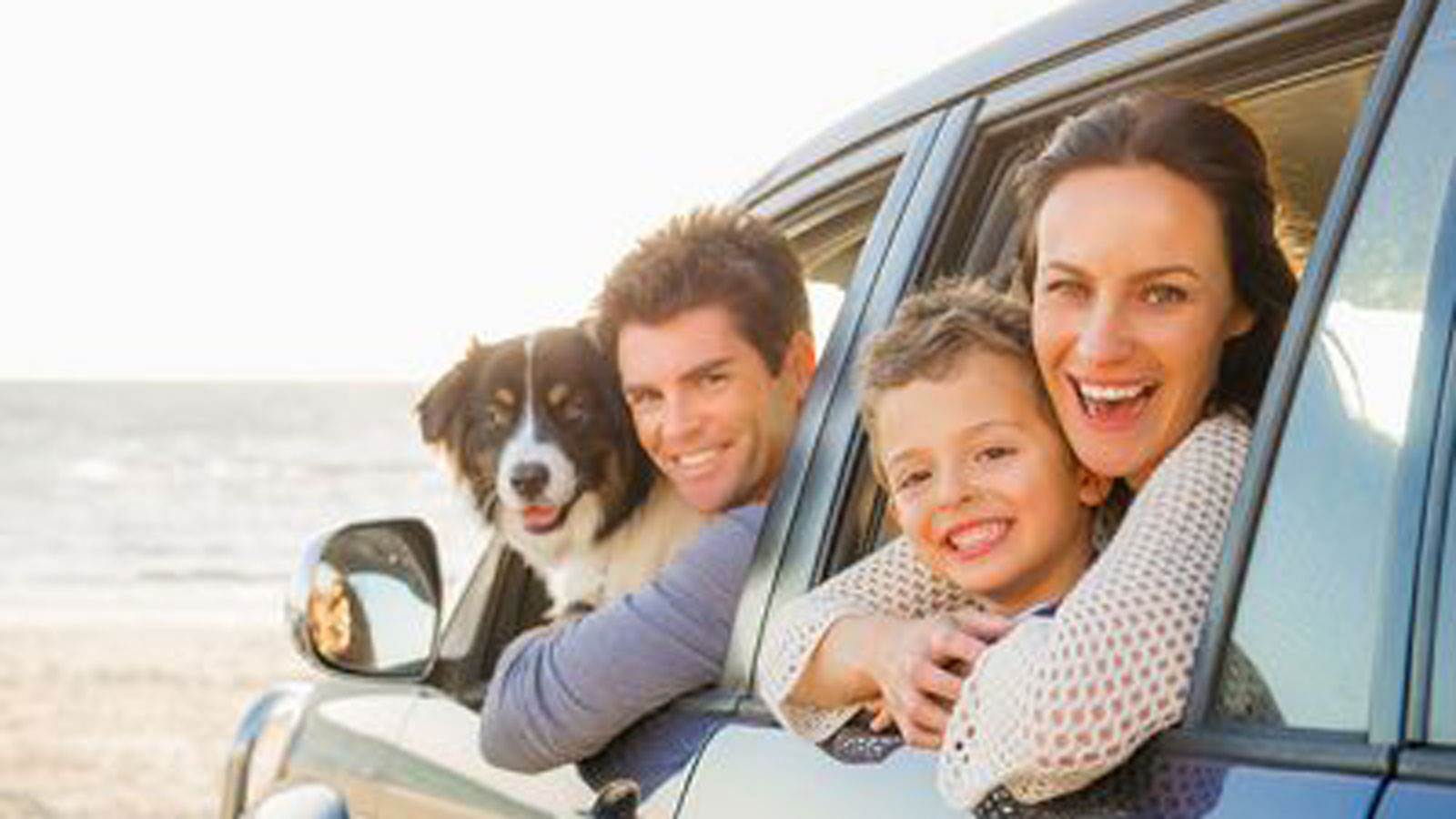 A young family and a dog smiling outside of a car window whilst at the beach.