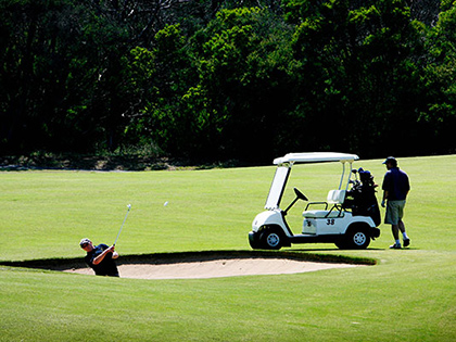 Person hitting a golf ball out of a sand bunker near a parked golf cart, at RACV Cape Schanck Resort.