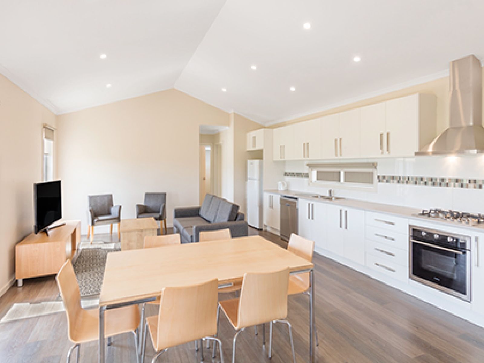 Living area and kitchen in RACV Cobram Resort's Garden View Cabin, featuring a long kitchen bench, six-seat dining table, couches and television.