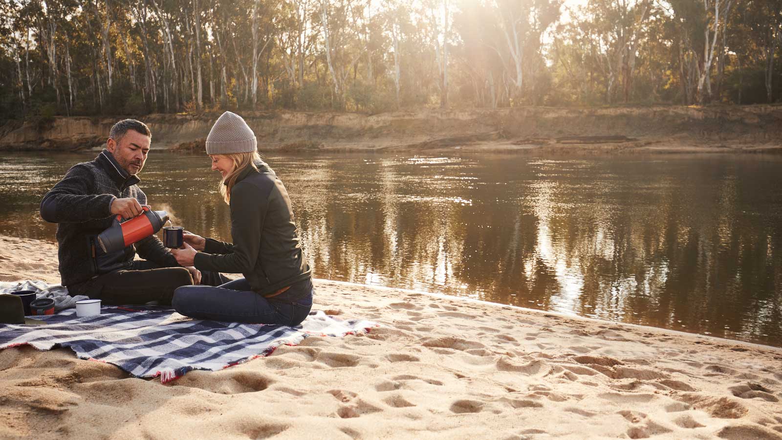 Couple seated on a picnic rug with thermos on the edge of the Murray River. 